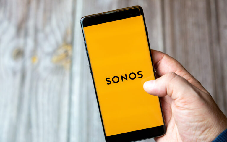 Eine Person hält ein Smartphone mit der Sonos-App in der Hand. Auf einem gelben Hintergrund mit dem Sonos-Logo in schwarzer Schrift wird ihr Soundsystem-Setup präsentiert. Die Holzoberfläche dahinter verleiht dem Ganzen einen Hauch von Wärme, während sie ihre Systemeinstellungen überprüft und das Telefon mit der rechten Hand hält.