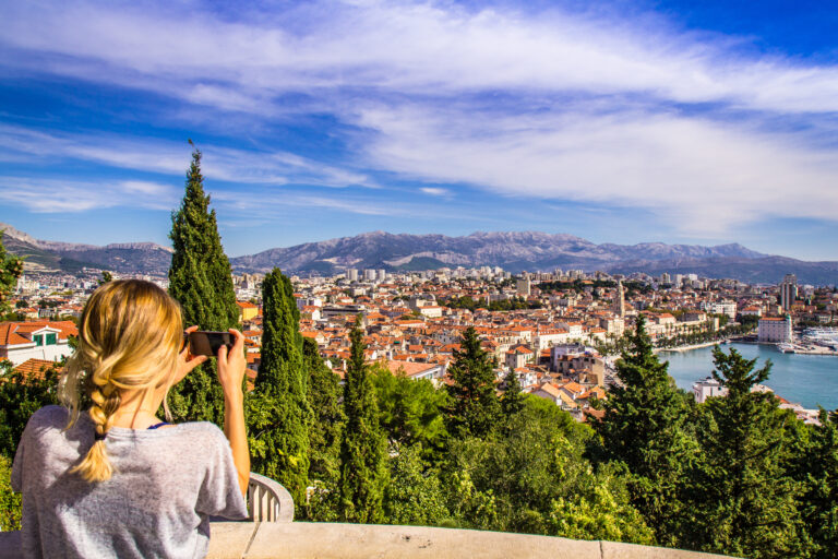 Eine Person mit blondem Haar hält ein Smartphone in der Hand und macht ein Foto von einer malerischen Aussicht auf eine Küstenstadt mit roten Dächern, üppigem Grün und bergigem Gelände unter einem klaren blauen Himmel. Das lebhafte und weitläufige Stadtbild scheint perfekt, um positive Bewertungen zu erhalten.