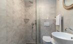 Ein modernes Badezimmer mit ebenerdiger Dusche, abgetrennt durch eine Glaswand, mit wandmontiertem Duschkopf, Chromarmaturen und eingebautem Regal. Daneben befindet sich eine Toilette mit Spülknopf an der Wand, einem runden Waschbecken und einem Spiegel darüber. Der Raum hat geflieste Wände.