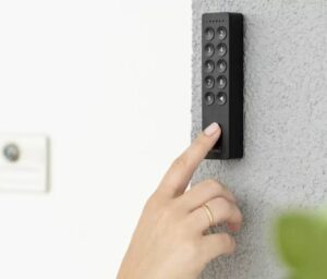 Eine Hand mit einem Ring am Zeigefinger drückt eine Taste auf einem schwarzen Tastenfeld, das an einer strukturierten Wand angebracht ist. Das Tastenfeld, das für einen einfacheren CheckIn unerlässlich ist, besteht aus Zahlen von 1 bis 9, die in einem Raster angeordnet sind. Im Hintergrund sind eine verschwommene Steckdose und im Vordergrund eine grüne Pflanze zu sehen.
