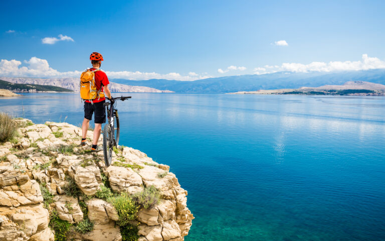 Ein Radfahrer in einem roten Hemd, einem orangefarbenen Helm und einem orangefarbenen Rucksack steht neben einem Mountainbike auf einer Felsklippe und blickt auf einen ruhigen blauen See mit Bergen in der Ferne unter einem klaren Himmel.